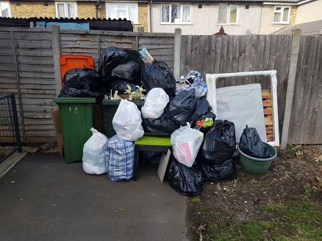 A collection of black, white, and patterned rubbish bags are piled on and around a green bench against a wooden fence in an outdoor area, likely a backyard or alleyway. The bags contain household waste and are stacked on the bench, with some leaning against the wooden fence and others placed on the ground. To the left of the rubbish, there is a large green waste bin with a closed lid, and behind the pile, an orange recycling container is partially visible. On the right side, there is a large, empty white picture frame or panel leaning against the fence, and beside it, a green plastic planter with no visible plants inside. The ground is paved with asphalt, and there is a small patch of soil with sparse grass or weeds next to the fence. The scene suggests a typical rubbish disposal situation, and it is relevant to waste removal services provided by Commercial Waste Weybridge, situated near the Weybridge area.