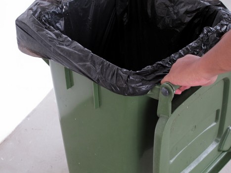 A close-up view of a green wheeled rubbish bin with a black plastic liner partially pulled back, revealing a dark interior indicating it is used for waste disposal. The bin is positioned on a concrete surface, with a person's hand visible gripping the handle of the lid, opening or closing it. The background is plain and neutral, emphasizing the bin's sturdy plastic construction and the textured surface of the waste liner. This scene illustrates typical waste collection equipment used by Commercial Waste Weybridge for local rubbish removal services, often situated on driveways or outside commercial or residential premises in Weybridge or nearby areas. The metallic handle on the lid provides ease of use, and the overall setup suggests readiness for rubbish disposal, aligning with professional waste management practices in the region.