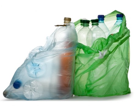 A collection of clear plastic bottles, including varying sizes and colors such as blue, green, and transparent, are shown inside and around a partially transparent green plastic bag that is loosely folded. The bottles appear to have caps, with some caps remaining on and others removed, indicating they were previously used for beverages. The bottles and bag are placed on a plain, light-colored surface with a white background, emphasizing the materials' glossy and smooth textures. The scene is evenly lit, highlighting the reflective surfaces of the bottles and the semi-sheen finish of the plastic bag. This arrangement demonstrates typical waste items suitable for collection and removal services, such as those offered by Commercial Waste Weybridge, located near Weybridge, Surrey. The visual focus on recyclable plastic bottles aligns with local rubbish removal or recycling initiatives, providing a neutral and factual depiction of waste ready for disposal or recycling processes.