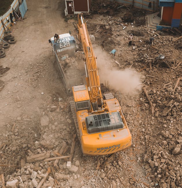 An aerial view of a construction site showing a large, orange excavator with a long boom arm and bucket attachment, actively demolishing a section of a building or structure. The excavator has visible